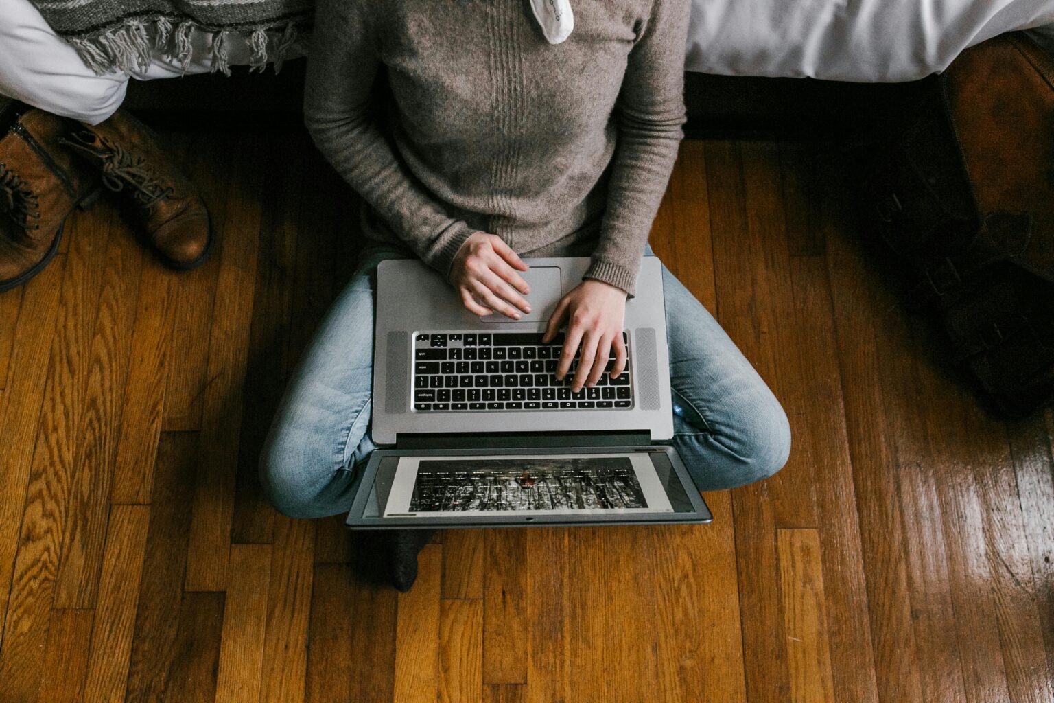 A young woman sitting on a hardwood floor working on a laptop in a modern bedroom setting.